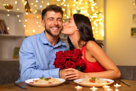 Foto de Alegre cena romántica como mujer sonriente en vestido rojo besa amorosamente la mejilla de un hombre encantado sosteniendo rosas, celebrando un momento íntimo - Imagen libre de derechos