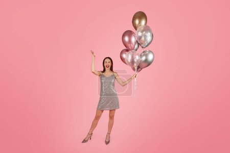 Téléchargez les photos : Femme millénaire caucasienne exubérante avec un large sourire, levant joyeusement la main, tenant une grappe de ballons métalliques, d'humeur festive lors d'une fête avec un fond rose - en image libre de droit