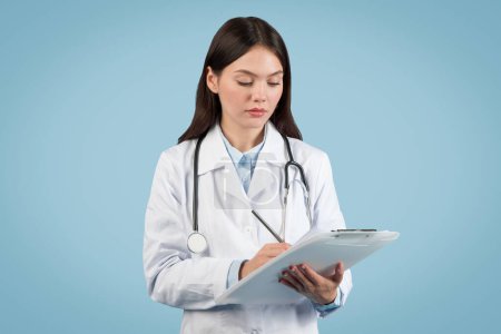 Téléchargez les photos : Jeune femme médecin concentrée en blouse de laboratoire prenant des notes sur presse-papiers, avec stéthoscope autour du cou, sur fond bleu propre - en image libre de droit