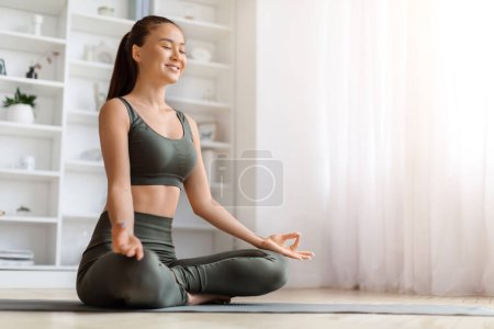 Foto de Hermosa mujer asiática joven sonriendo suavemente mientras practica yoga en casa, mujer coreana tranquila meditando en la estera en la sala de estar, realizando mudra, encarnando relajación y alegría, espacio para copiar - Imagen libre de derechos