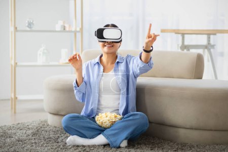 Téléchargez les photos : Femme asiatique excitée utilisant un casque VR à la maison, assise sur le sol dans un salon confortable, touchant monsieur, regardant un film, mangeant du pop-corn, faisant l'expérience de la réalité virtuelle - en image libre de droit