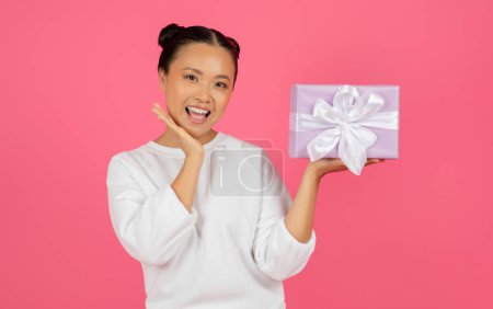 Aufgeregt fröhlich asiatische Frau hält Geschenkbox auf ihrer Handfläche, überrascht junge Koreanerin zeigt Geschenk und anrührendes Gesicht in Aufregung, steht isoliert auf rosa Studiohintergrund