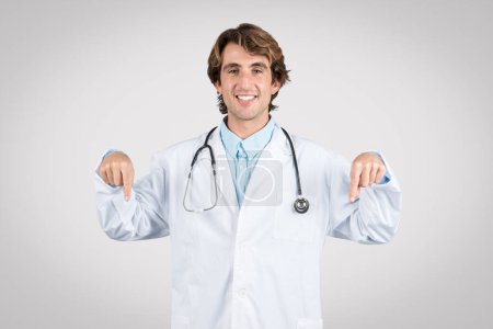 Téléchargez les photos : Médecin masculin joyeux en blouse blanche pointe vers le bas avec les deux mains, souriant, indiquant éventuellement des informations sur la santé ou la zone de concentration ci-dessous - en image libre de droit