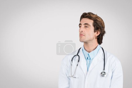 Photo for Confident male doctor wearing white coat and stethoscope gazes at free space, symbolizing professionalism and contemplation in healthcare, grey background - Royalty Free Image