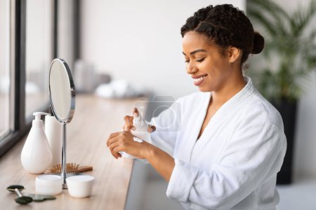 Téléchargez les photos : Femme noire souriante utilisant la crème hydratante pour les mains à la maison, Jeune femme afro-américaine portant un peignoir blanc frottant lotion corporelle nourrissante à la peau, assis près du miroir dans la salle de bain - en image libre de droit