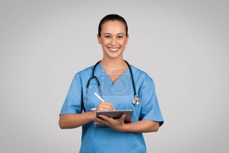 Infirmière joyeuse en blouse bleue utilisant une tablette numérique, avec stéthoscope autour du cou, symbolisant la technologie de soins de santé moderne, fond gris
