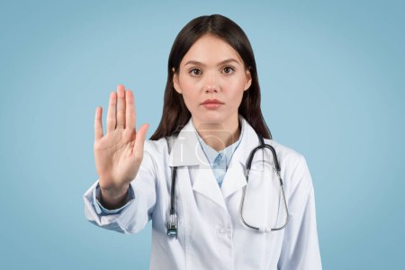 Ernsthafte Ärztin mit Stethoskop um den Hals macht Stoppgeste, die Vorsicht oder Ablehnung im Gesundheitswesen anzeigt, auf blauem Hintergrund