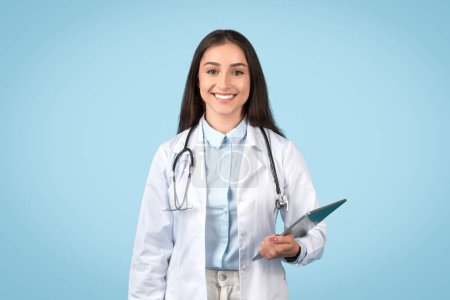 Téléchargez les photos : Médecin professionnel tenant presse-papiers prêt pour les notes du patient, avec un sourire agréable et portant un blouse blanche, sur fond bleu tranquille - en image libre de droit