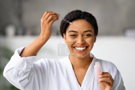 Foto de Cuidado de belleza. Sonriente atractiva mujer negra aplicando suero facial con gotero mientras hace rutina de cuidado de la piel en casa, hermosa mujer afroamericana hidratante piel y mirando a la cámara - Imagen libre de derechos