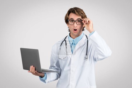 Téléchargez les photos : Choqué médecin masculin ajuster ses lunettes, tenant ordinateur portable avec expression perplexe, peut-être réagir à des données médicales inattendues ou des nouvelles, fond gris - en image libre de droit