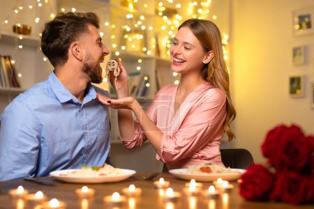 Téléchargez les photos : Homme heureux sur le point de goûter spaghetti ludique offert par la main des femmes, les deux partageant un moment joyeux lors d'un dîner aux chandelles avec des roses. - en image libre de droit