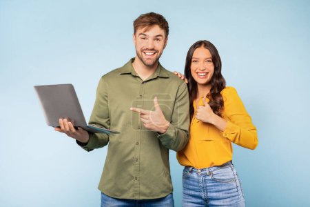Téléchargez les photos : Joyeux homme et femme avec ordinateur portable, homme pointant vers l'ordinateur tandis que la femme donne pouces vers le haut, indiquant l'approbation ou le succès sur fond bleu vif - en image libre de droit