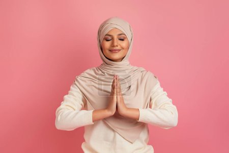 Foto de Mujer musulmana árabe alegre en hijab de pie con los ojos cerrados y las manos apretadas, encarnando la serenidad y la gratitud, posando sobre fondo de estudio rosa, simbolizando la armonía interior y el orgullo cultural - Imagen libre de derechos