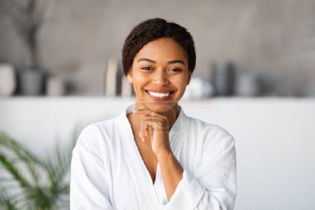 Photo for Beautiful young black woman wearing white bathrobe touching face and smiling at camera, attractive african american female enjoying relaxing day at home or at modern, serene spa setting, copy space - Royalty Free Image