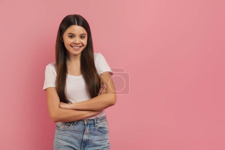 Photo for Confident teen girl smiling standing with her arms crossed and looking at camera, positive smiling female teenager dressed in white t-shirt and jeans standing on pink studio background , copy space - Royalty Free Image