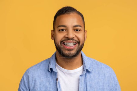 Téléchargez les photos : Joyeux jeune homme noir avec des appareils dentaires portant chemise bleue posant en toute confiance sur un fond jaune optimiste, beau mec afro-américain exsudant positivité et style, espace de copie - en image libre de droit