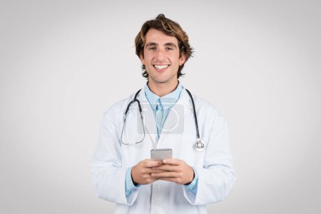Feliz médico masculino de bata blanca con estetoscopio mensajes de texto en el teléfono inteligente, que representa el uso de aplicaciones móviles en la atención médica moderna, contra el fondo gris