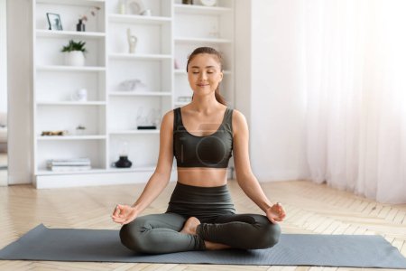 Téléchargez les photos : Serene jeune femme asiatique en vêtements de sport pratiquant la méditation avec les yeux fermés, calme coréen femme assise en position lotus sur tapis de yoga dans lumineux, minimaliste salon à la maison, espace de copie - en image libre de droit