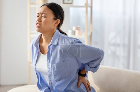 Photo for Sad millennial chinese woman rubbing lower back, sitting on couch at home, have pain or muscle strain after uncomfortable pose. Backache reasons, copy space - Royalty Free Image