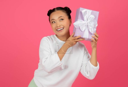 Adivina qué hay dentro. Mujer asiática sorprendida escuchando lo que traquetea en caja de regalo, mujer coreana positiva sacudiendo presente, pensando y sonriendo, de pie aislado sobre fondo de estudio rosa, espacio libre