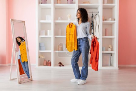 Téléchargez les photos : Femme chic en jeans contemplant des choix de mode, tenant chemise jaune et rouge, avec son reflet visible dans un miroir pleine longueur, dans une pièce rangée - en image libre de droit