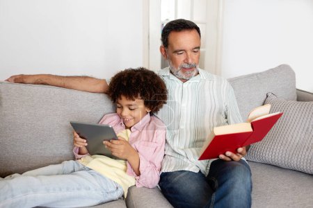 Foto de Ocio Familiar. Hombre mayor hispano leyendo libro mientras su nieto usa la tableta digital, relajándose juntos en el sofá en la sala de estar moderna en casa. Internet y pasatiempo tradicional - Imagen libre de derechos