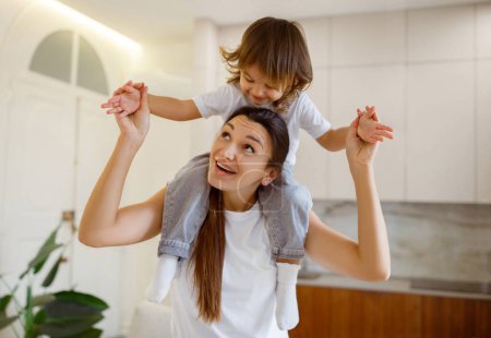 Photo for Joyful young mother giving her laughing daughter piggyback ride, happy mom and cute little female child playing together with cheerful expressions in bright and airy home environment - Royalty Free Image