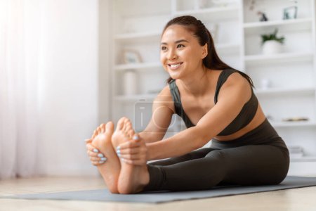 Téléchargez les photos : Joyeuse femme asiatique faisant assis vers l'avant plier l'exercice d'étirement tout en s'entraînant à la maison, belle jeune coréenne pratiquant le yoga sur tapis dans le salon, profiter de l'entraînement domestique, copier l'espace - en image libre de droit