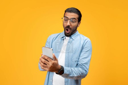 Photo for A startled man in a blue shirt stares at his smartphone with wide eyes and open mouth, showing a reaction of shock or surprise at a message or notification, against a bright yellow background. - Royalty Free Image