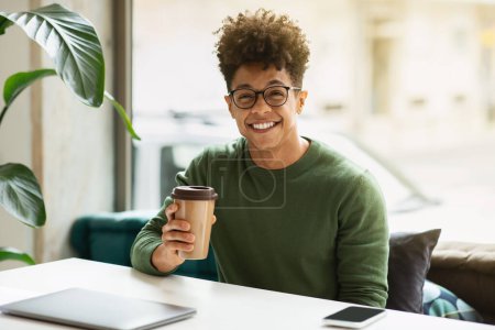 Téléchargez les photos : Élégant homme afro-américain aux cheveux bouclés portant des lunettes freelancer boire du café dans un café confortable, assis seul à table par la fenêtre, souriant à la caméra. Loisirs, concept de pause café - en image libre de droit