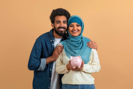 Foto de Feliz pareja musulmana abrazando y sosteniendo alcancía, sonriendo jóvenes cónyuges árabes mostrando la responsabilidad financiera y los objetivos de ahorro, de pie contra el fondo beige estudio, espacio de copia - Imagen libre de derechos