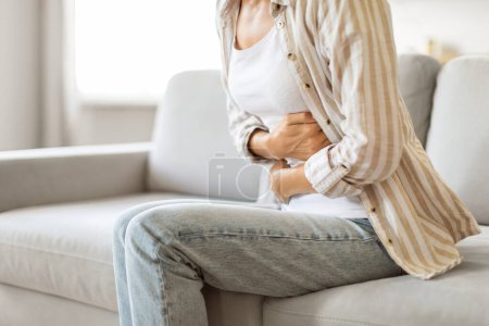 Photo for Uncomfortable young woman holding her abdomen, feeling unwell with stomach ache while sitting on couch in well-lit living room, unrecognizable lady suffering digestion problems, cropped shot - Royalty Free Image