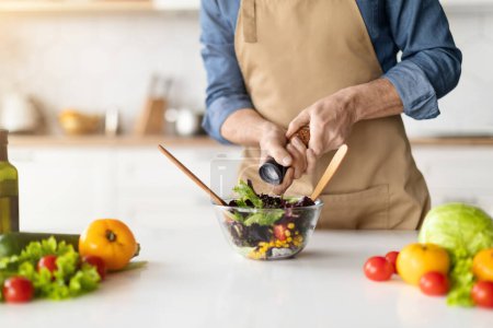 Photo for Man in apron seasoning colorful bowl of salad with fresh vegetables spread on the kitchen counter, unrecognizable male showcasing healthy home cooking, adding salt or pepper to meal, closeup - Royalty Free Image