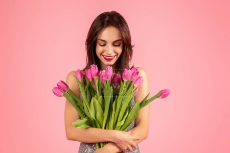 Photo for Admiring woman in a sequined dress smiles gently at a large bouquet of pink tulips shes embracing, a moment of happiness and serenity on a pink backdrop - Royalty Free Image