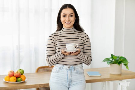 Foto de Mujer joven sonriente que presenta una taza de café con ambas manos, de pie en una cocina con luz natural y un tazón de fruta fresca sobre la mesa. Pausa para el café, bebida de rutina - Imagen libre de derechos