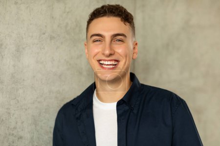 A joyful glad caucasian young man with a beaming smile, wearing a navy blue shirt and a white t-shirt, against a concrete textured background. Work, study and positive lifestyle