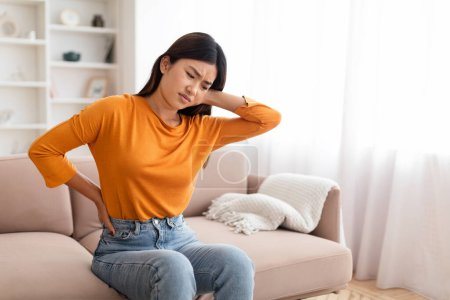 Photo for Sad millennial asian woman sitting on couch in cozy living room at home, touching lower back and neck, feeling pain after nap on sofa, suffering from office syndrome after work, copy space - Royalty Free Image