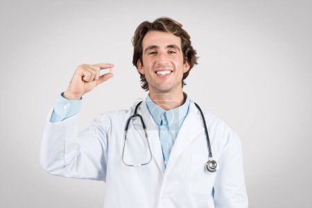 Lächelnder Arzt im Laborkittel mit Pille zwischen den Fingern, die das Konzept von Medikamenten oder Nahrungsergänzungsmitteln illustriert, grauer Hintergrund