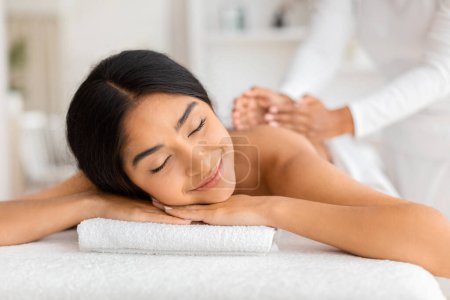 Téléchargez les photos : Joyeuse jeune femme indienne recevant un massage relaxant du dos par un thérapeute professionnel dans un salon de spa, souriante jolie dame détendue bénéficiant d'un traitement de bien-être dans un salon de spa - en image libre de droit