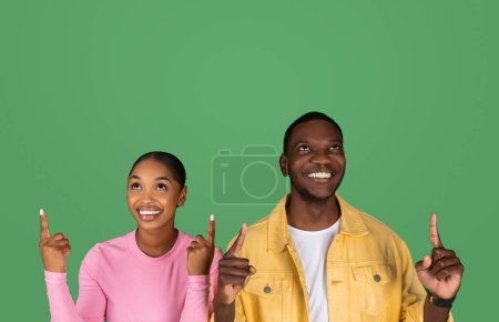 Photo for Happy millennial black couple checking great offer, pointing up at copy space for advertisement above their heads, isolated on green studio background. Great deal for two - Royalty Free Image