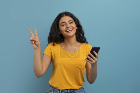 Photo for Nice mobile app, online offer. Positive attractive curly young hindu lady with smartphone in her hand showing peace gesture and smiling, blue studio background - Royalty Free Image