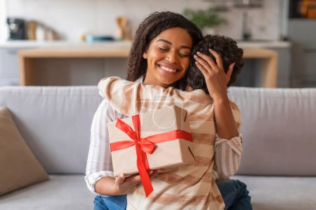 Photo for Joyful black mother holding wrapped gift box and affectionately embracing her son in gratitude, happy african american family of two sharing loving moment in their cozy living room, free space - Royalty Free Image
