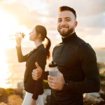 Energetic couple takes break on coastal path, man smiles with thumbs up and water bottle, woman hydrates against the backdrop of glowing sea horizon