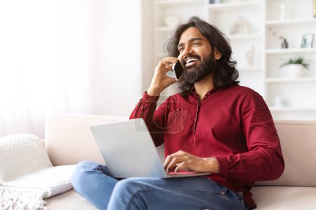 Téléchargez les photos : Barbu aux cheveux longs Indien à la recherche d'un emploi en ligne, assis sur le canapé avec ordinateur sur ses genoux, parlant au téléphone avec l'employeur, regardant l'espace de copie pour la publicité. Opportunités de carrière - en image libre de droit