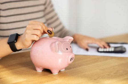 Photo for Hand is inserting a coin into a pink piggy bank, indicative of saving money, with a calculator and financial documents in the background, symbolizing budgeting for future - Royalty Free Image