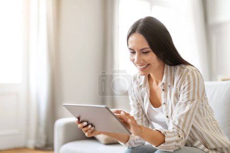 Photo for Cheerful young woman engaged with her digital tablet, sitting on comfortable sofa in bright living room interior, happy millennial female embodying relaxed tech-friendly lifestyle, resting at home - Royalty Free Image