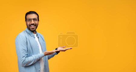 Foto de Un hombre sonriente con barba y gafas sostiene su mano plana y extendida, como si presentara un producto o característica, con una mirada de invitación amistosa, sobre un fondo naranja brillante - Imagen libre de derechos