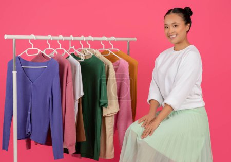 Belle jeune femme asiatique assise gracieusement près du porte-vêtements et souriant à la caméra, heureuse coréenne femme contemplant des choix de mode en boutique, posant sur fond de studio rose, gros plan