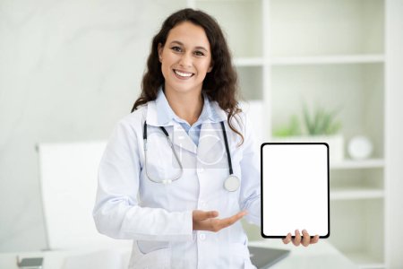 Téléchargez les photos : Joyeux médecin européen millénaire femme tenant tablette numérique avec écran blanc espace de copie de maquette, recommandant application mobile pour les technologies modernes dans les soins de santé, intérieur de la clinique - en image libre de droit