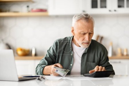 Photo for Concentrated senior male with cash in hand calculating finances at home, elderly gentleman sitting at kitchen table with laptop and documents, making budget management and financial planning - Royalty Free Image